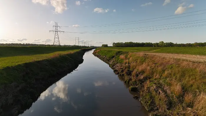 Calm river reflecting sky, flanked by grassy banks and power lines under a partly cloudy sky.