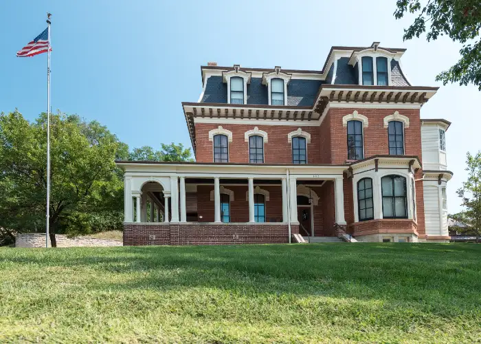 Three-story historic red brick mansion with white trim, wraparound porch, and American flag on pole.