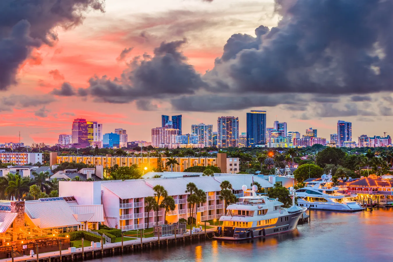 City skyline at sunset with high-rise buildings, yachts, waterfront, and dramatic clouds.