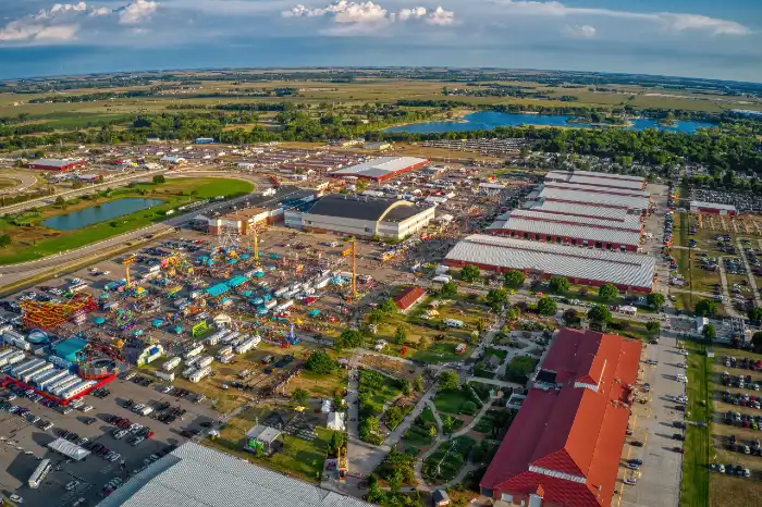 Aerial view of a large outdoor fairground with colorful rides, tents, and surrounding buildings under a partly cloudy sky.