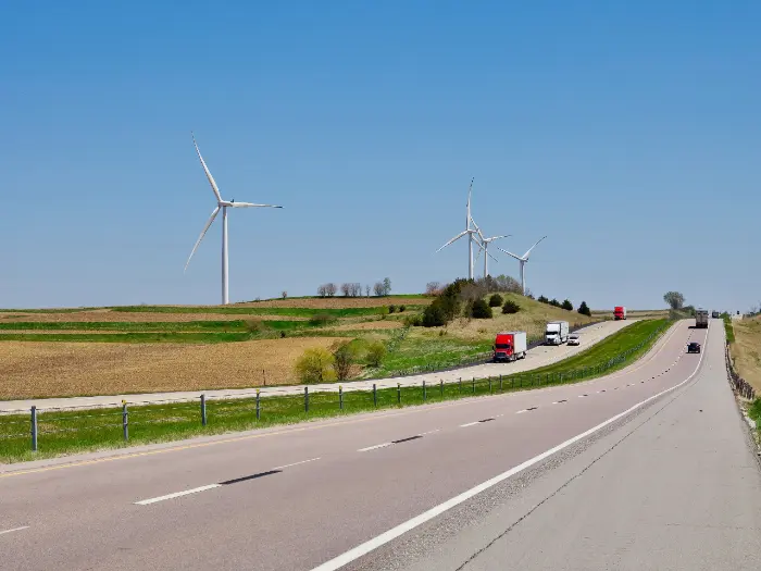Highway with multiple trucks driving, adjacent green fields, and three large wind turbines under clear blue sky.