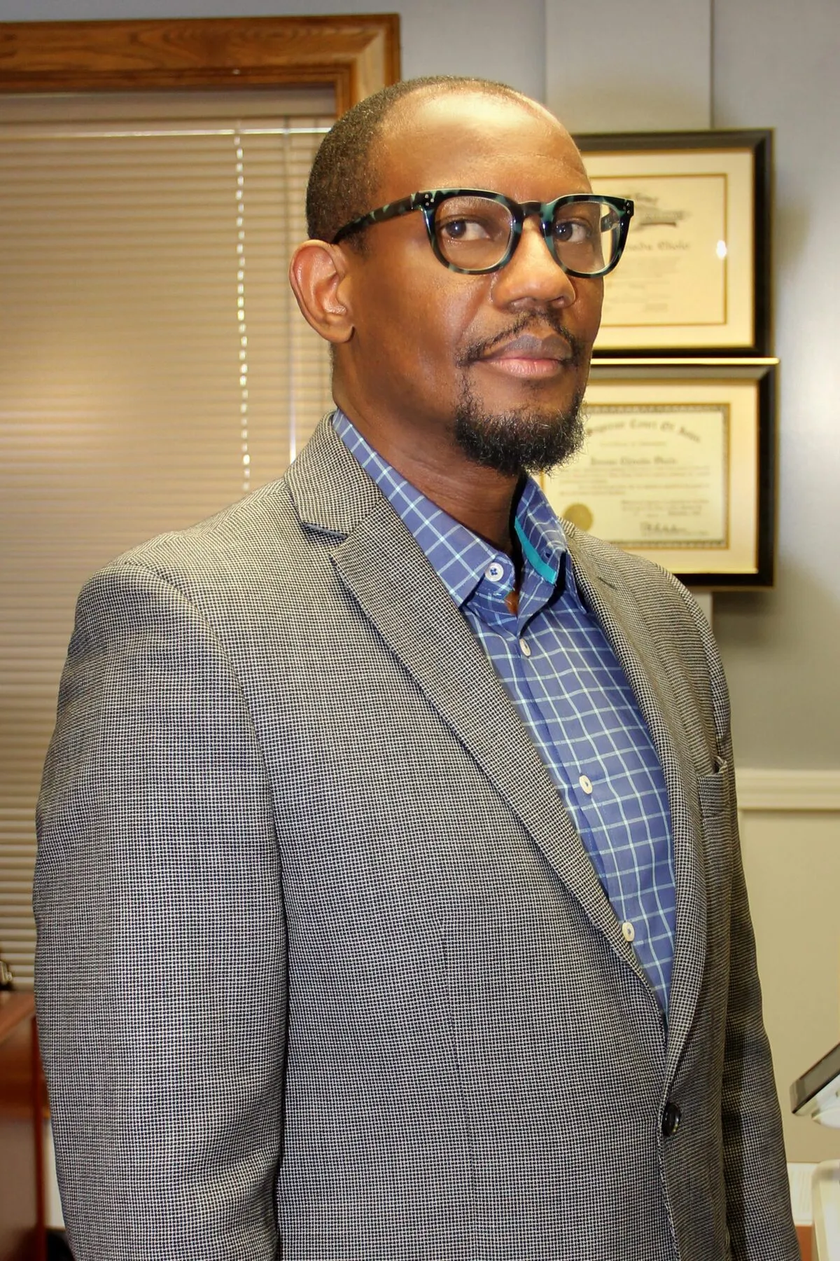 Professional Black man wearing tortoiseshell glasses, gray checked blazer, and blue checkered shirt, standing in office.