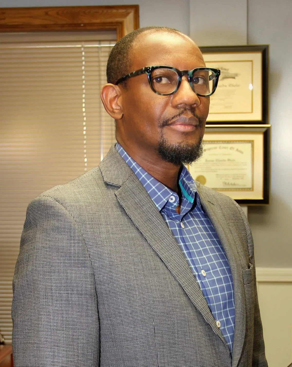 Professional man wearing glasses, gray blazer, and blue checkered shirt, standing in office with framed certificates.