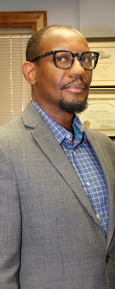 Professional man wearing glasses, gray checked blazer, and blue checkered shirt, standing indoors with framed certificates in background.