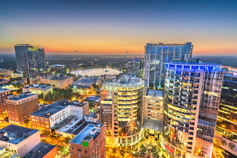 Aerial view of a cityscape at sunset with illuminated high-rise buildings and a distant lake.