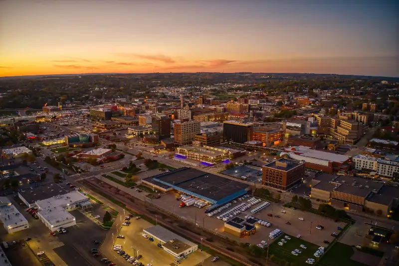 Aerial view of a cityscape at sunset with illuminated buildings and parking lots.