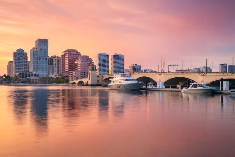 City skyline with modern buildings, boats docked by a bridge at sunset.