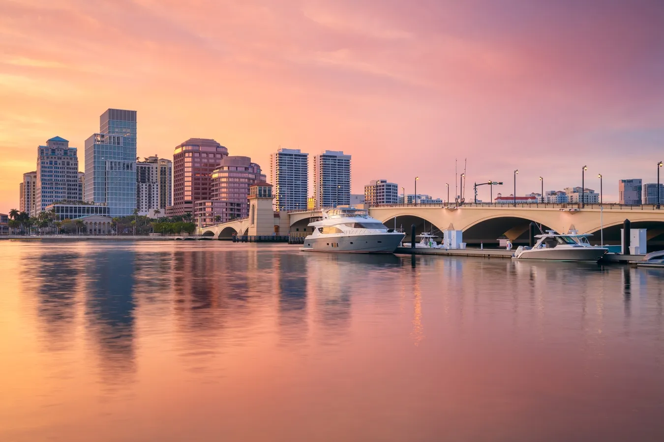 Calm waterfront with docked yachts, arched bridge, and modern city skyline at sunset.