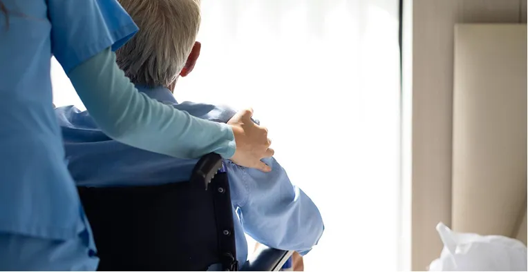Healthcare professional in blue scrubs comforting elderly person in wheelchair by window.
