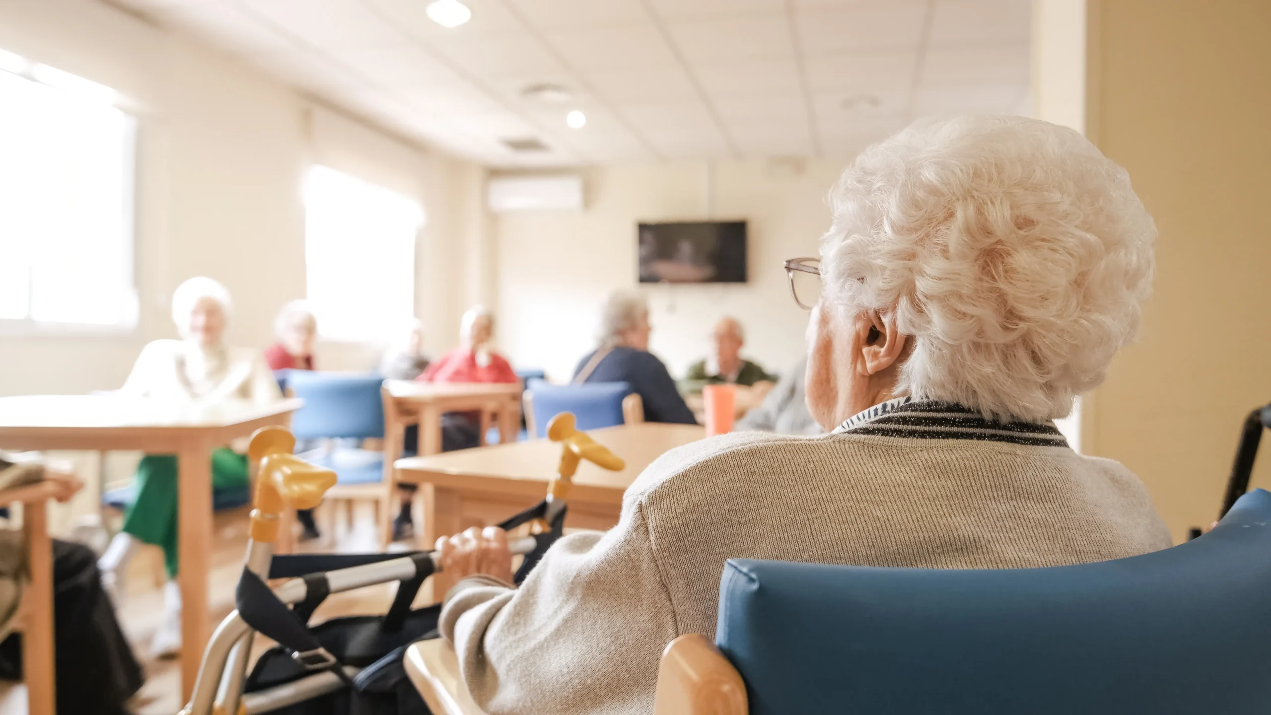 Elderly individuals seated in a bright common room of a senior care facility.