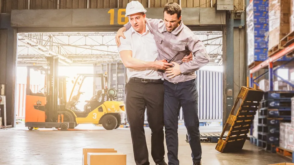 Two men in a warehouse, one wearing a white hard hat supporting an injured man clutching his side.