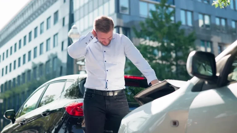 Man in white shirt standing between two cars after a minor rear-end collision in an urban setting.
