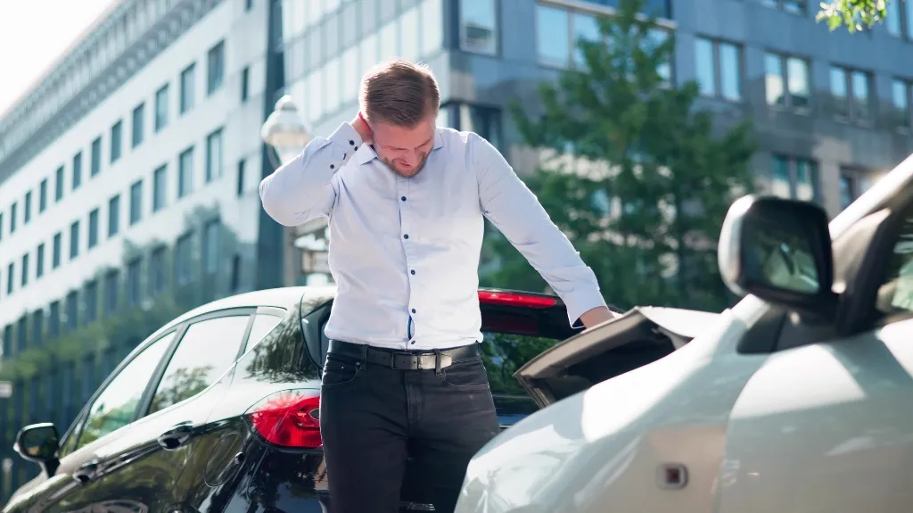 Man in white shirt standing between two cars after a minor rear-end collision in an urban setting.