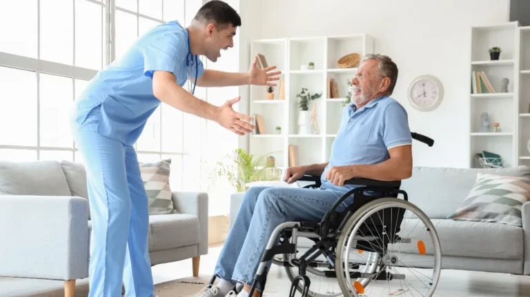 Healthcare professional in blue scrubs engaging positively with elderly man in wheelchair indoors.