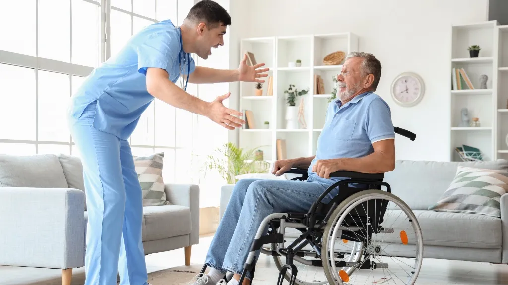 Healthcare professional in blue scrubs engaging positively with elderly man in wheelchair indoors.