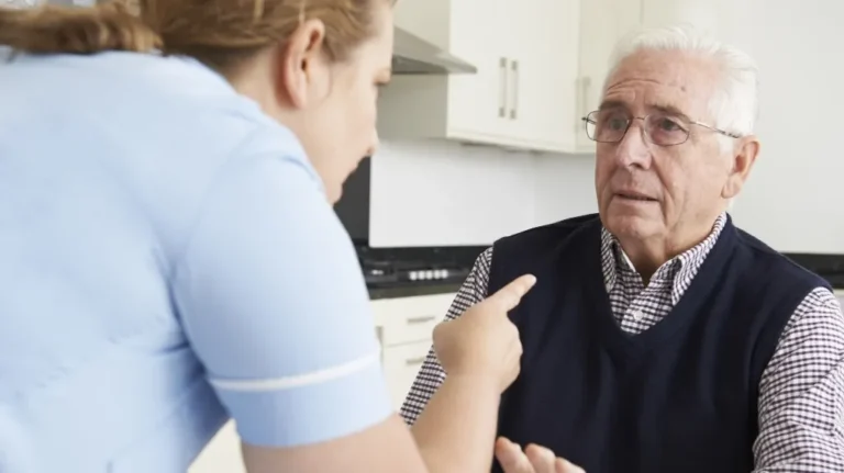Healthcare professional in blue uniform speaking to concerned elderly man with white hair and glasses.