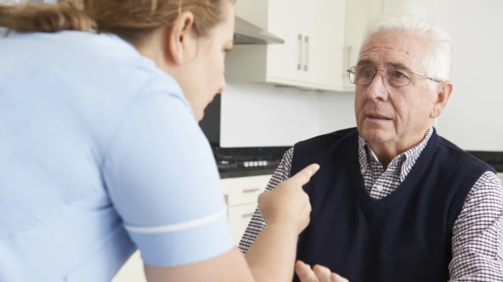Healthcare professional in blue uniform speaking to concerned elderly man with white hair and glasses.