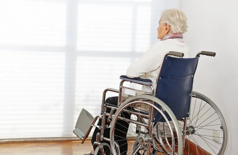 Elderly person with white hair seated in a blue wheelchair, facing a bright window.