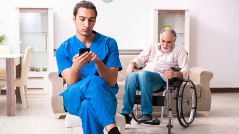 Young male healthcare worker in blue scrubs using smartphone, elderly man in wheelchair in background.